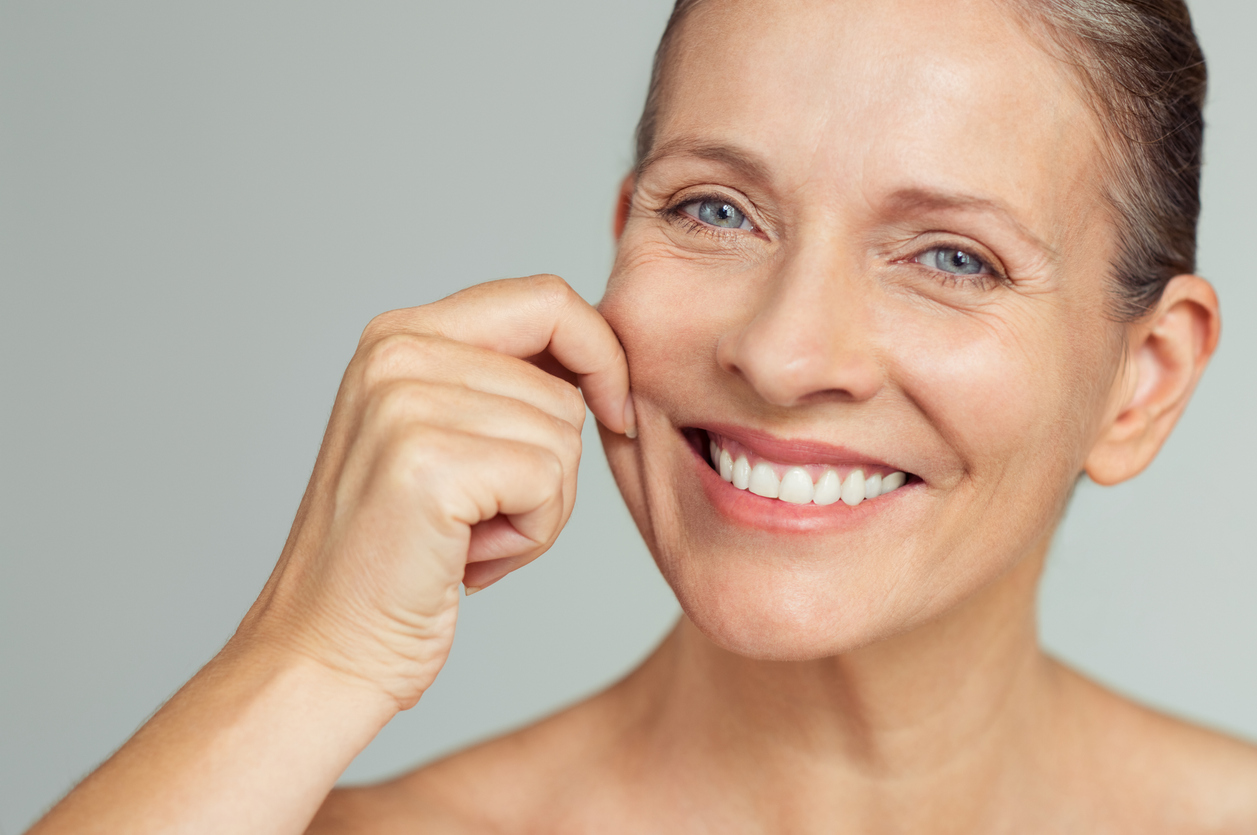 Senior woman pulling cheeks to feel softness and looking at camera. Beauty portrait of happy mature woman smiling with hands on cheek isolated over grey background. Aging process and perfect skin concept.